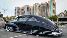 an old black car parked in a parking lot next to tall buildings and palm trees