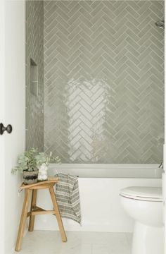 a white toilet sitting next to a bath tub under a window in a bathroom with gray tiles on the walls