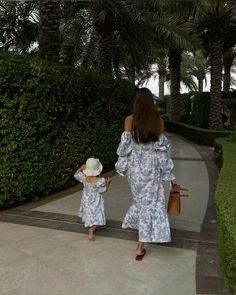 a woman and child walking down a sidewalk next to some bushes with palm trees in the background