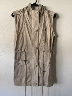 "Vintage Zenana Outfitters Sleeveless Utility Coat This item is brand new in perfect condition. Tag on item reads size \"S\". Please see item measurements listed below. 100% Cotton.  Safari Vibes! Beige sleeveless coat with two big fully functional snap closure pockets on front of garment. Zipper pocket on left chest side is decorative. Snap and zipper main closure. Tie on waste so you can cinch the garment for a more fitted look. Two snap military style shoulder tabs (decorative). Hood is rounded on top. Item Measurements (Garment Lying Flat) Shoulder - 16\" Bust - (Armpit to Armpit) 17\" Waist - 16\" Hem Width - 18\" Length - 27.5\" (Shoulder to Hem) Length - 37\" (Hood to Hem Hood Width - 12\" Hood Length - 10\" Item Care Dry Clean Only Made in Vietnam 🇻🇳  Item Condition Perfect Condi Utility Sleeveless Tops With Pockets, Utility Sleeveless Vest For Summer, Sleeveless Utility Top With Pockets, Summer Utility Sleeveless Vest, Summer Sleeveless Utility Vest, Casual Sleeveless Khaki Vest, Casual Khaki Sleeveless Vest, Sleeveless Khaki Outerwear For Spring, Khaki Sleeveless Outerwear For Spring