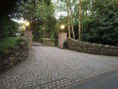 a brick driveway surrounded by stone walls and gates with two lamps on each side of the gate