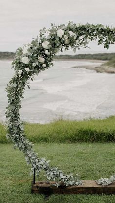 a wedding arch with white flowers and greenery on the grass by the water in front of an ocean