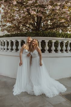 two women in white dresses standing next to each other near a tree with pink flowers