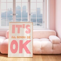 a pink couch sitting next to a window in a living room under a poster that says, it's all gonna be ok