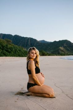 a pregnant woman sitting on the beach in a black bathing suit with mountains in the background