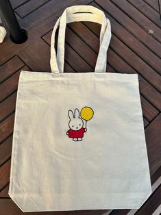 "A canvas tote bag with an embroidered miffy character with a yellow balloon  Dimensions: 15\"L x 16\"H 9 1/2\" handle drop 100% Cotton" Cute Tote Bags Paint, Miffy Embroidery, Embroidered Tote Bag Ideas, Miffy Items, Totebag Painting, Painted Totes, Embroidered Ideas, Tote Bag Inspo