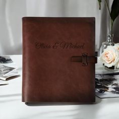 a brown leather guest book on a table with flowers and photos in the back ground