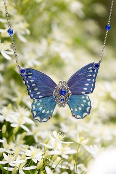 a blue butterfly necklace is hanging from a chain with white flowers in the foreground