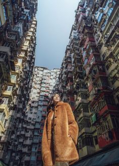 a woman wearing a fur coat standing in front of tall buildings with balconies