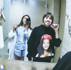 two young women brushing their teeth in front of a bathroom mirror while another woman takes a selfie