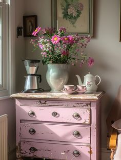 a pink dresser with flowers in a vase and two tea kettles on top of it