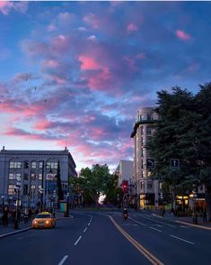 a car driving down a street next to tall buildings at dusk with pink clouds in the sky