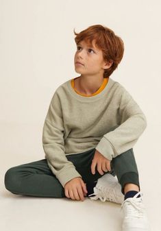 a young boy sitting on the ground wearing green pants and a grey sweater with an orange stripe