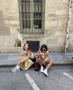 two young women sitting on the ground with their cell phones in front of an old building