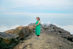 a woman in a green dress standing on rocks near the ocean with her arms crossed