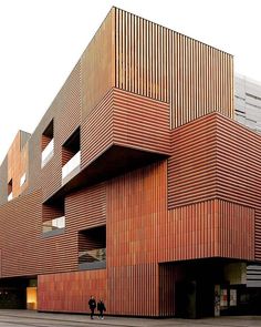 two people walking in front of a building with wooden slats on the facade and sides