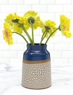 yellow flowers in a blue vase on a marble countertop with white tiles behind it