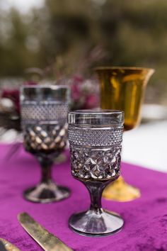 three glass goblets sitting on top of a purple table cloth next to silverware