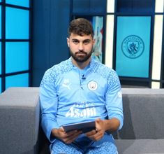 a man sitting on top of a gray couch next to a blue wall holding a tablet