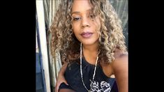 a woman with long curly hair wearing a black tank top and pearls necklaces, standing in front of a window