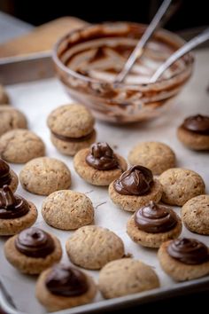 cookies with chocolate frosting are on a baking sheet next to a bowl of peanut butter