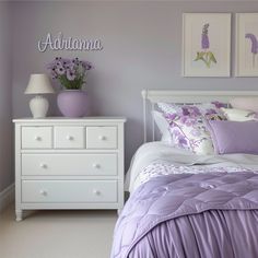 a white bed with purple comforter and pillows in a bedroom next to two pictures on the wall