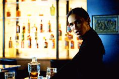 a man sitting at a table with two glasses in front of him and bottles on the wall behind him