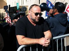 a man in black shirt and sunglasses standing next to a fence with people behind him