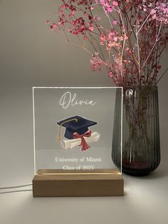 a clear acrylic plaque with a graduation cap and tassel on it next to a vase filled with pink flowers