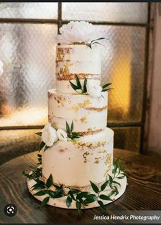 a three tiered cake with white flowers and greenery sits on a table in front of a window