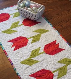 a quilted table runner with red and green leaves on it, next to a white basket filled with eggs