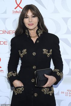 a woman in black coat and gold trims holding a clutch on the red carpet