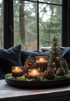 candles are arranged on a tray with pine cones and evergreens in front of a window