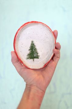 a hand holding a small ceramic christmas ornament with a green tree on it