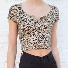 a woman with short hair wearing a leopard print top and black pants, standing in front of a white brick wall