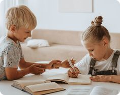 two young children sitting at a table with books and pens in their hands, looking at an open book