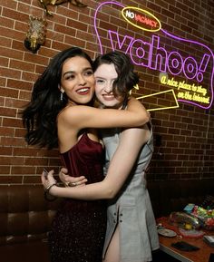 two young women hugging each other in front of a brick wall with neon signs behind them