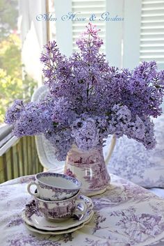 purple flowers are in a vase on a table next to two cups and saucers