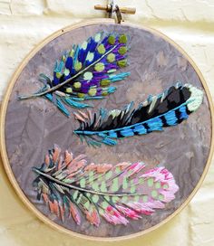 three different colored feathers are in a circular embroidery hoop on a white wall behind it