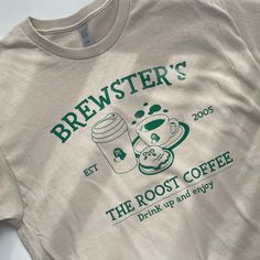 The Roost Coffee T-Shirt by petimint is a beige t-shirt with green text and graphics, featuring the words BREWSTER'S, EST 2005, THE ROOST COFFEE, Drink up and enjoy. The graphics showcase a to-go coffee cup, a steaming mug, and a plate with a pastry—making it an ideal piece of apparel for any gamer inspired by Stardew Valley. Coffee Shop T Shirt Design, Cafe T Shirt Design, Tshirt Design Minimal, Vintage Style Tshirts, Cafe Shirt Design, T Shirt Design Inspiration Graphic Tees Retro Vintage, Coffee Shop Shirts, Fun Shirt Designs, Coffee Shop Tshirt Design