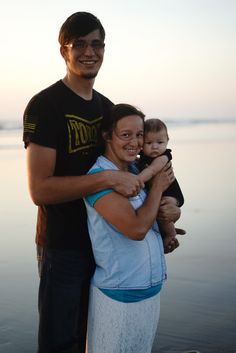 a man and woman holding a baby standing on the beach at sunset with water in the background