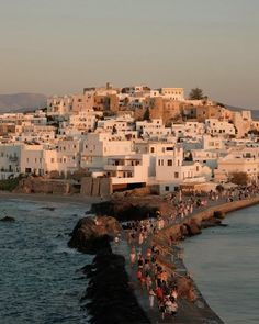 many people are walking along the water near some houses and buildings on top of a hill