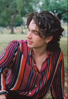 a young man sitting on the ground in a park wearing a striped shirt and earrings