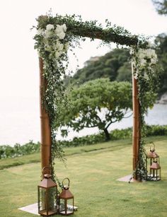 an outdoor ceremony setup with lanterns, flowers and greenery on the grass by the water