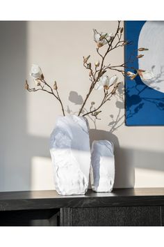 two white vases with flowers in them on a table next to a painting and wall