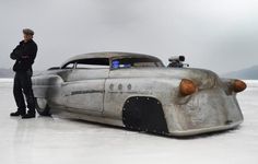 a man standing next to an old car in the middle of a frozen lake with mountains in the background