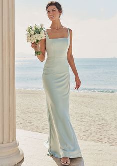 a woman in a long dress standing on the beach holding flowers and smiling at the camera