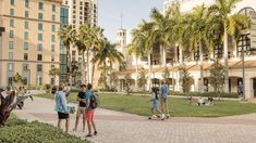 several people are standing on the sidewalk in front of a building with palm trees and tall buildings