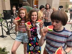 three young children eating watermelon slices in front of a group of people on the sidewalk