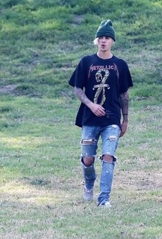 a young man in ripped jeans and a green hat walks through the grass with his skateboard